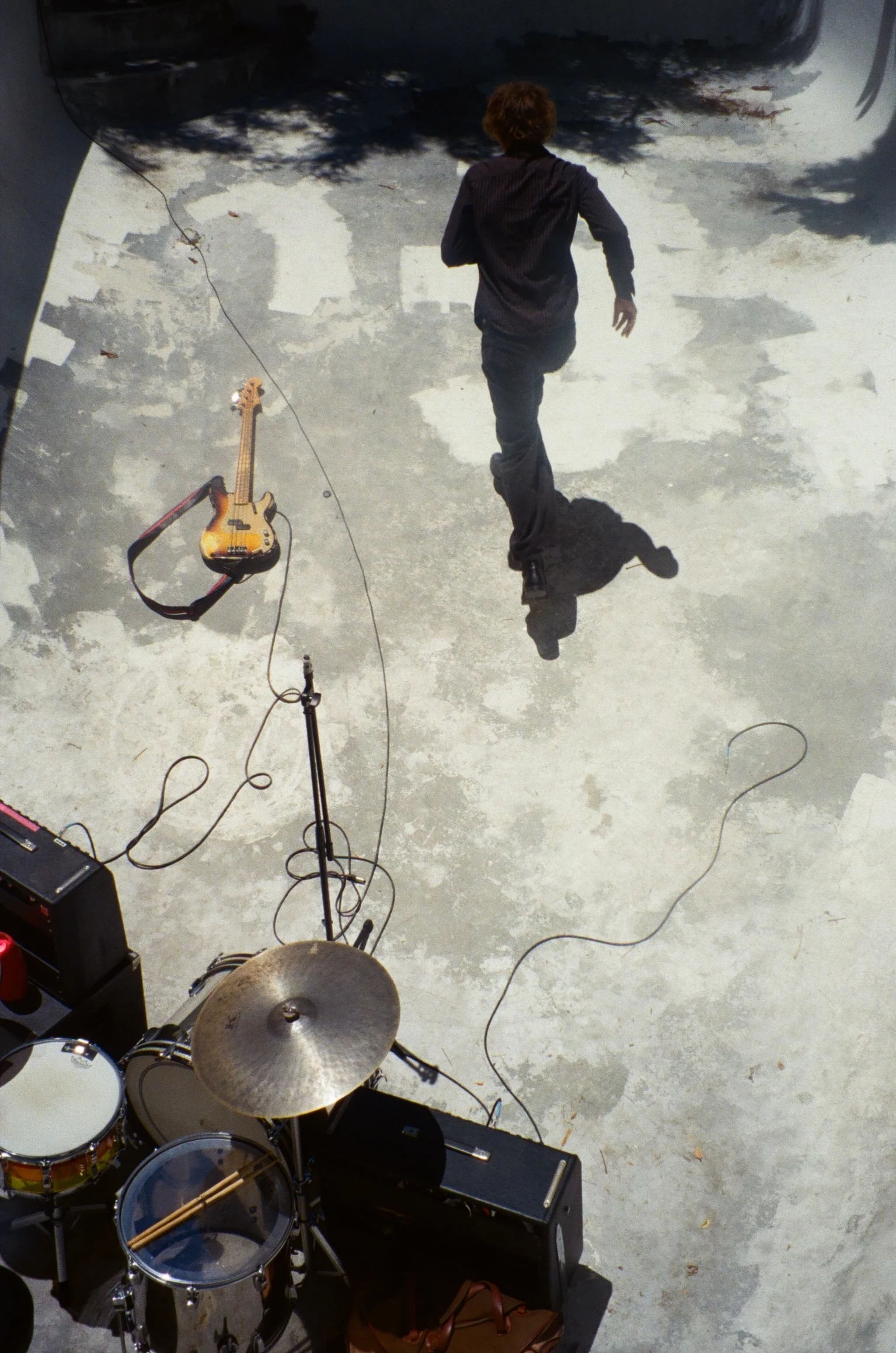 A musician crossing a sunlit stage beside a bass guitar and drum kit.
